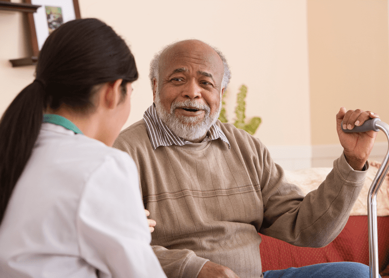 Smiling elderly man holding a cane, engaging in conversation with a healthcare professional during an in-home visit.