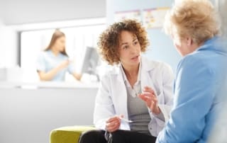 Female doctor having a focused conversation with an older patient in a clinic setting, with another healthcare worker in the background.