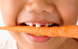 Close-up of a child smiling while biting on a whole carrot, showing missing front teeth.